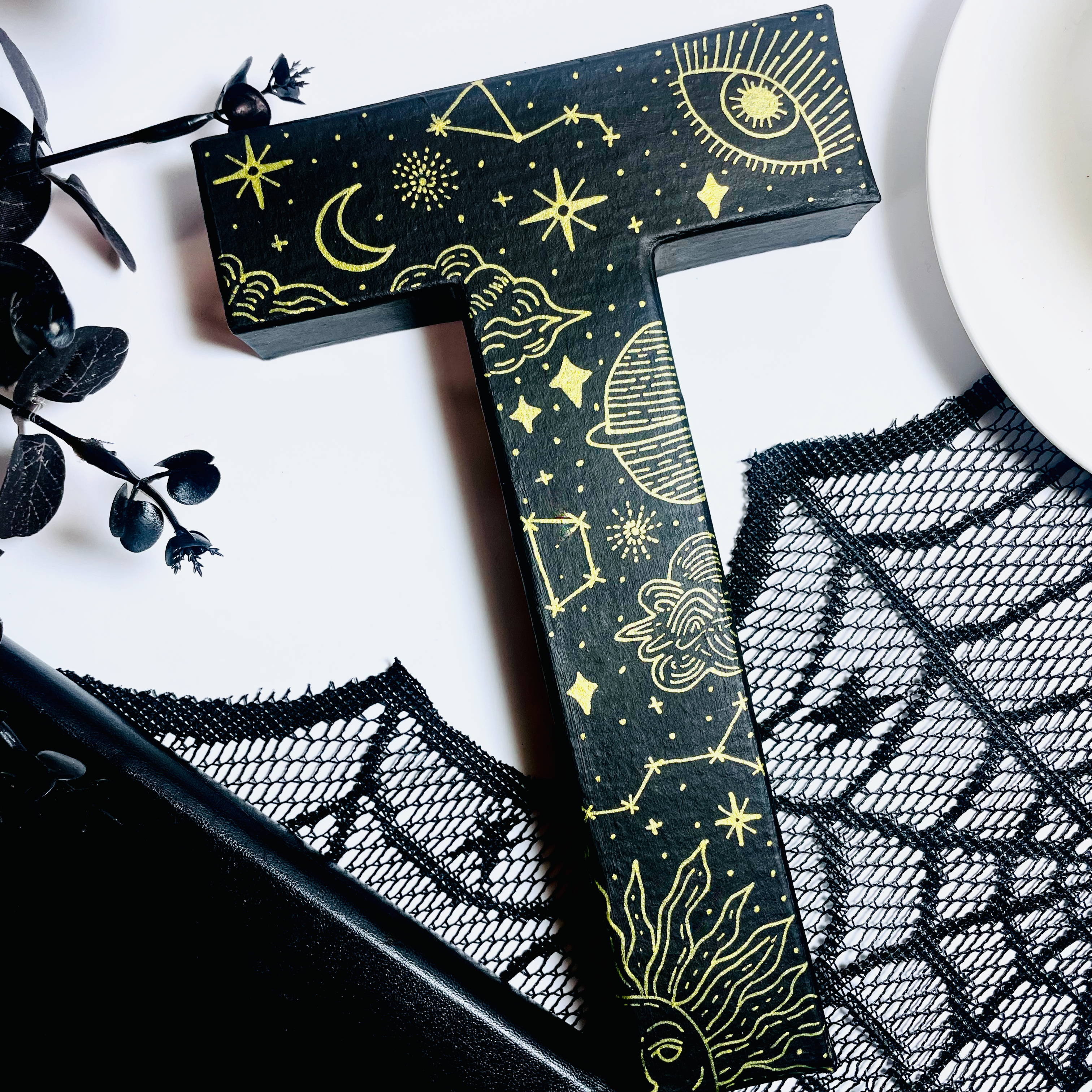 Decorative letter 'T' with celestial designs on a white surface with black lace and flowers.