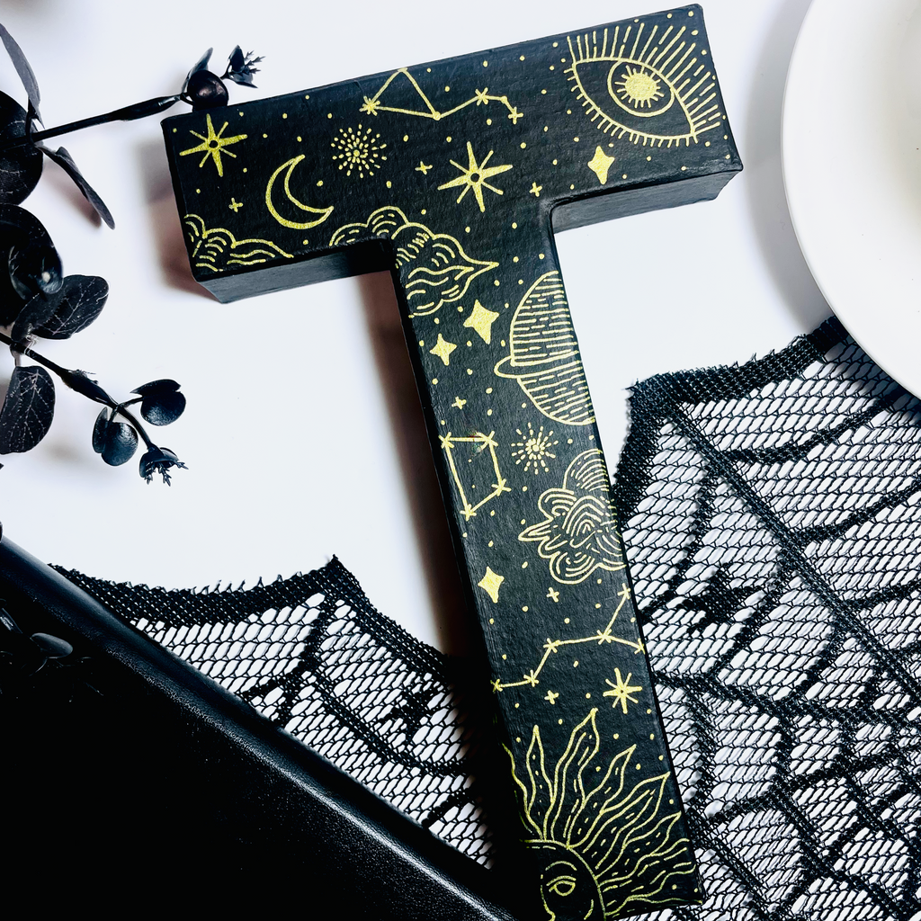Decorative letter 'T' with celestial designs on a white surface with black lace and flowers.