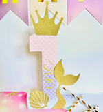 Load image into Gallery viewer, A mermaid-themed first birthday party decoration set featuring a large glittery gold number 1 with a mermaid tail and seashell design, gold crown, and striped paper straws on a pastel pink and white background.
