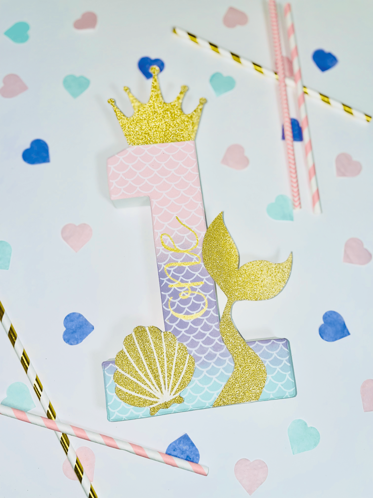 Mermaid themed number '1' with a gold glitter crown, tail and shell on a white background with pastel confetti hearts scattered along with white and gold straws for a themed display.